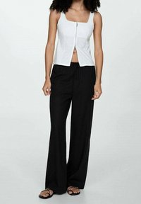 Débardeur zippé blanc à encolure carrée associé à un pantalon large noir. Le haut présente un design ajusté et une texture douce. Des sandales noires complètent la tenue.