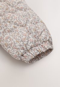 Quiltad tygärm med elastisk mudd, med ett litet blommönster i mjuk rosa, beige och grått på vit bakgrund.