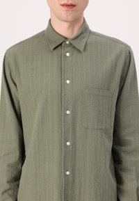 Camicia a maniche lunghe verde, con texture, colletto button-down, bottoni frontali e taschino sul petto. Il tessuto presenta un motivo sottile.