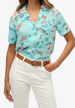 Vrouw draagt een turquoise Hawaiiaans overhemd met korte mouwen en een bloemen- en gitaarprint, ingestopt in een witte broek met een bruine riem.