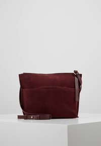 Burgundská semišová kabelka s plochým obdélníkovým tvarem, nastavitelným koženým popruhem a přední kapsou. Hladká textura s minimálními kovovými prvky.