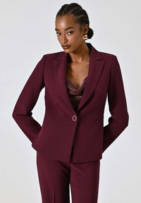 Blazer aderente color bordeaux con revers a lancia, chiusura con un bottone e due tasche frontali. Abbinato a un pantalone coordinato. Tessuto con texture liscia.