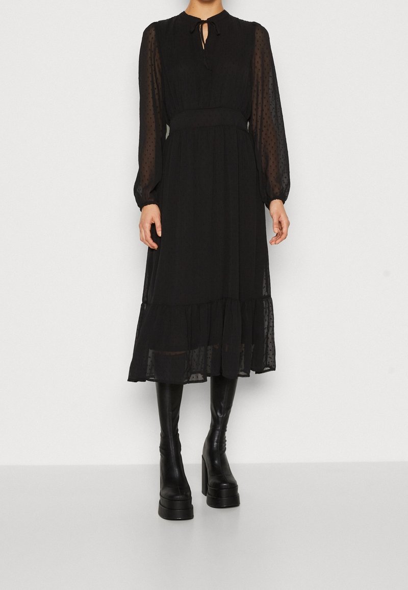 Femme portant une robe midi noire avec des manches longues transparentes, des poignets froncés, un décolleté en forme de goutte et des bottes noires à plateforme montant jusqu'aux genoux.