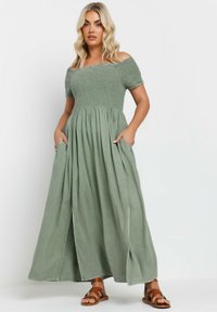 Maxime dress i mjuk grön tyg, med av-axeln ärmar, smockad bodice, fickor och en flödande kjol med sidofickor.
