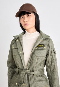 Olijfgroene gewatteerde jas met grote kraag, voorzakken en tailleband. Gecombineerd met een bruine pet en een witte blouse met ruches.