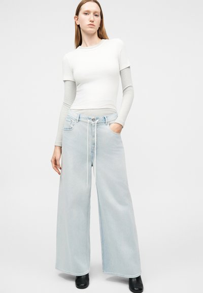 Γυναίκα στέκεται φορώντας λευκό και γκρι στρωματοποιημένο μακρυμάνικο τοπ, ανοιχτό μπλε τζιν παντελόνι με φαρδιά μπατζάκια και κορδόνι, και μαύρα μποτάκια στον αστράγαλο σε απλό φόντο.