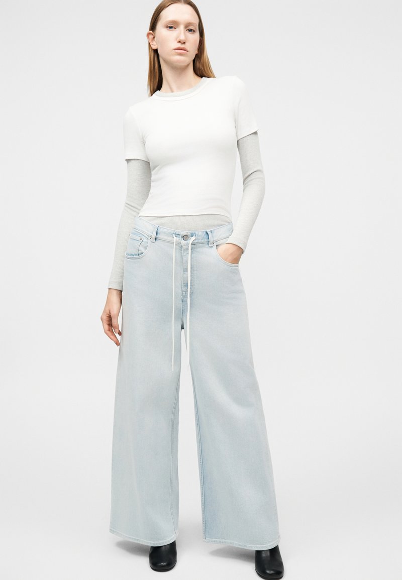 Woman stands wearing layered white and gray long-sleeve top, light blue wide-leg jeans with drawstring, and black ankle boots on plain background.