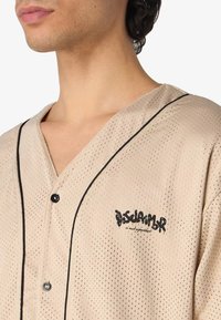 Camicetta in rete beige con bordi neri, maniche corte e chiusura a bottoni. Presenta un logo stampato sul petto. Texture perforata per traspirabilità.