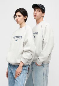 Kaksi nuorta aikuista seisoo vierekkäin päällään vaaleanharmaat New Balancen urheilupaidat ja vaaleansiniset farkut valkoisella taustalla.