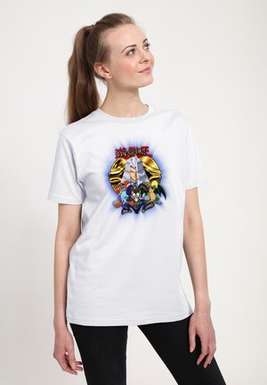 Ung kvinde med ponytail iført en hvid Yu-Gi-Oh T-shirt med figurer og et gyldent Millennium Puzzle-symbol.