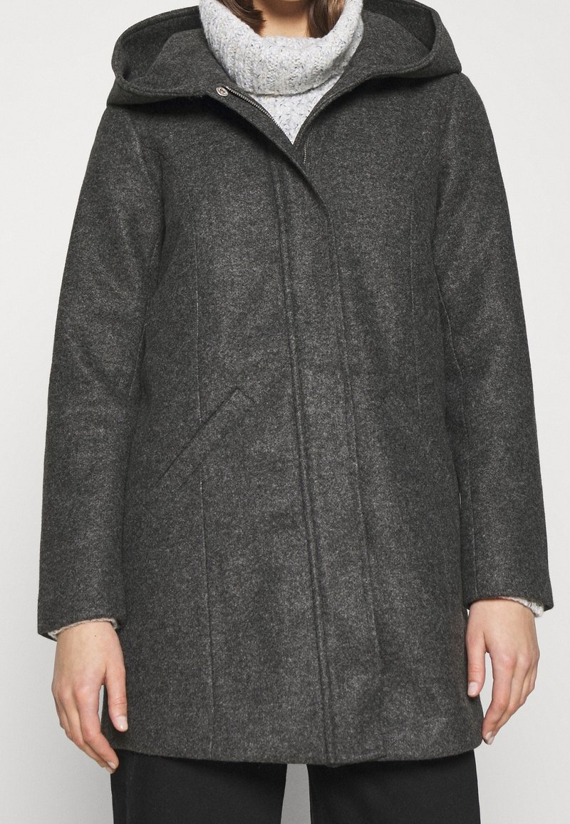 Personne portant un manteau en laine à capuche gris foncé sur un pull à col roulé en maille épaisse gris clair, debout devant un fond uni.