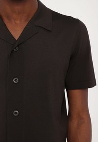 Chemise noire à manches courtes avec boutons et col en encoche, portée par une personne montrée du cou jusqu'au milieu du torse sur un fond uni.