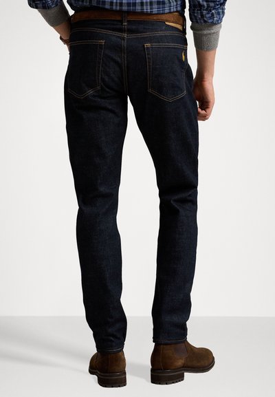 Jeans in denim blu scuro con cuciture arancioni a contrasto, slim fit, due tasche posteriori e una cintura in pelle marrone. Indossati con stivali alla caviglia marroni.