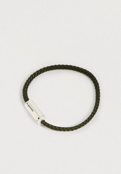Σκοτεινό πράσινο βραχιόλι από πλεγμένο κορδόνι με ασημένια μεταλλική κλείστρα, χαραγμένη με "le gramme", σε απλή λευκή φόντο.