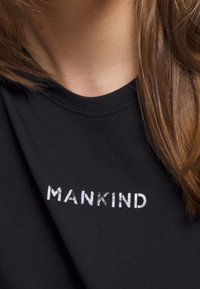 Nærbillede af en person iført en sort skjorte med hvid tekst, der står "MANKIND" nær kraven, som viser en del af deres hals og hår.