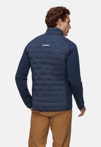 Mørkeblå jakke med polstret krop og glatte ærmer, med høj krave og brandnavnet "MAMMUT" på bagsiden.