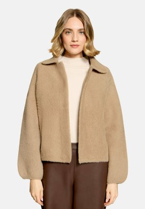 Vrouw met kort blond haar draagt een beige zachte jas over een crèmekleurige top en een bruine broek tegen een egale lichte achtergrond.