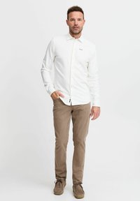 Cămașă albă cu mâneci lungi, cu deschidere la nasturi și trei puncte colorate pe piept, asortată cu chinos bej și pantofi din suede maron deschis.