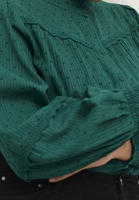 Groene textuur blouse met hoge hals, knoopsluiting en kanten accenten. Bevat verticale strepen en kleine stippenpatronen door het geheel.