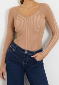 Beige gerippter Langarm-Body mit V-Ausschnitt, der strukturierte vertikale Streifen aufweist. Kombiniert mit dunkelblauen hochgeschnittenen Jeans.