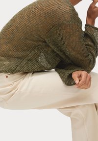 Maglione a maglia a rete verde oliva con maniche lunghe, indossato sopra pantaloni color crema. Il maglione presenta una vestibilità ampia e un motivo strutturato.