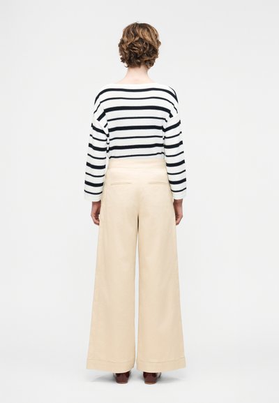 Άτομο με κοντά σγουρά μαλλιά που φοράει λευκό μακρυμάνικο μπλουζάκι με μαύρες ρίγες και μπεζ ψηλόμεσα παντελόνια φαρδιά, φαίνεται από την πίσω πλευρά.