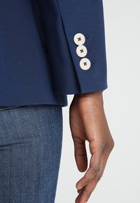 Sako námořnické modré barvy s rukávy, které mají čtyři světlé knoflíky, texturovanou tkaninu a dobře padnoucí střih, v kombinaci s džíny pro kontrast.