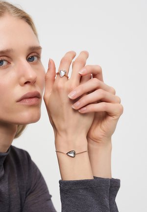 Frau mit heller Haut, die einen herzförmigen Ring an ihrem Finger und ein passendes herzförmiges Armband an ihrem Handgelenk trägt, die Hände sanft gefaltet.