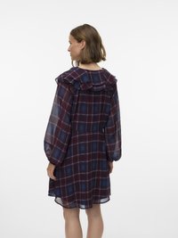 Sieviete, tērpusies caurspīdīgā tumši sarkanā un zilā pleda kleitā ar volānu apkakli un garām pūkainām piedurknēm, redzama no muguras uz balta fona.