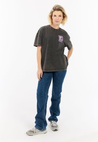 Grijze oversized t-shirt met een roze graphic op de linkerkant van de borst, gecombineerd met blauwe denim jeans en grijze sneakers. Glad stoftextuur.