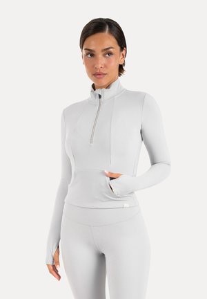 Ung kvinde iført lysegrå langærmet atletiktop med frontlynlås og matchende leggings, stående med den ene hånd i lommen.