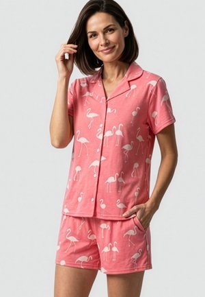 Vrouw draagt roze korte mouw gestikte blouse en short set met witte flamingo print, glimlachend met hand bij haar en andere in zak.