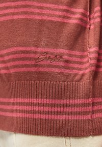 Κοντινό πλάνο στο στρίφωμα καφέ και ροζ ριγωτού πλεκτού πουλόβερ με ριμπ τελείωμα και κεντημένο όνομα μάρκας "Bugat" στο ύφασμα.