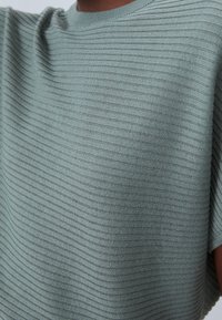 Zbliżenie na osobę w jasnozielonym swetrze o prążkowanym wzorze z poziomymi pasami i krótkimi rękawami.