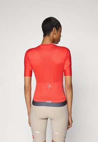 Jersey de ciclismo rojo de manga corta con tejido texturizado, bolsillos traseros y cierre con cremallera, combinado con pantalones cortos de ciclismo beige.