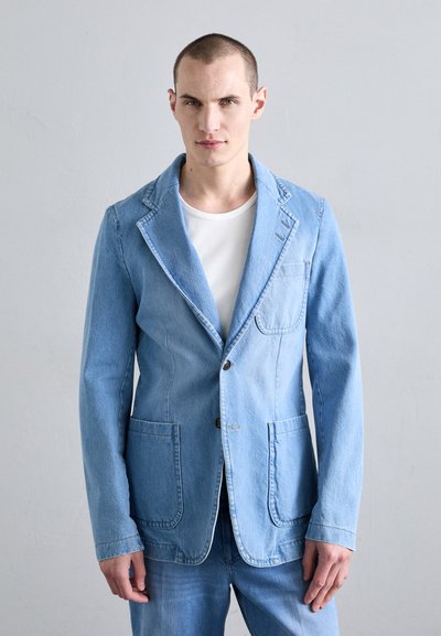 Denimblazer i lyseblå med klippet krave, to forlommer, knaplukning og skræddersyet pasform. Båret over en hvid T-shirt.