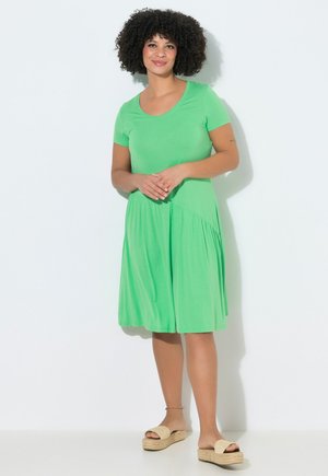 Frau mit lockigem Haar, die ein knielanges, knallgrünes Kleid mit kurzen Ärmeln und beigen Plateausandalen trägt, steht vor einer weißen Wand.