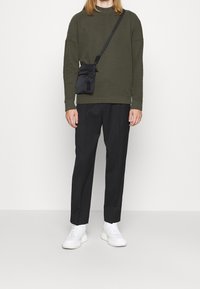 Camisa de manga larga color verde oliva, pantalones negros y zapatillas blancas. Bolsa cruzada negra con cremalleras. Tela suave y corte informal.