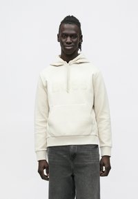 Νεαρός άνδρας με σκουρόχρωμο δέρμα και πλεγμένα μαλλιά, που φοράει ανοιχτό μπεζ φούτερ με κουκούλα με την επιγραφή "BOSS" και σκούρα γκρι τζιν, στέκεται μπροστά σε ένα απλό φόντο.