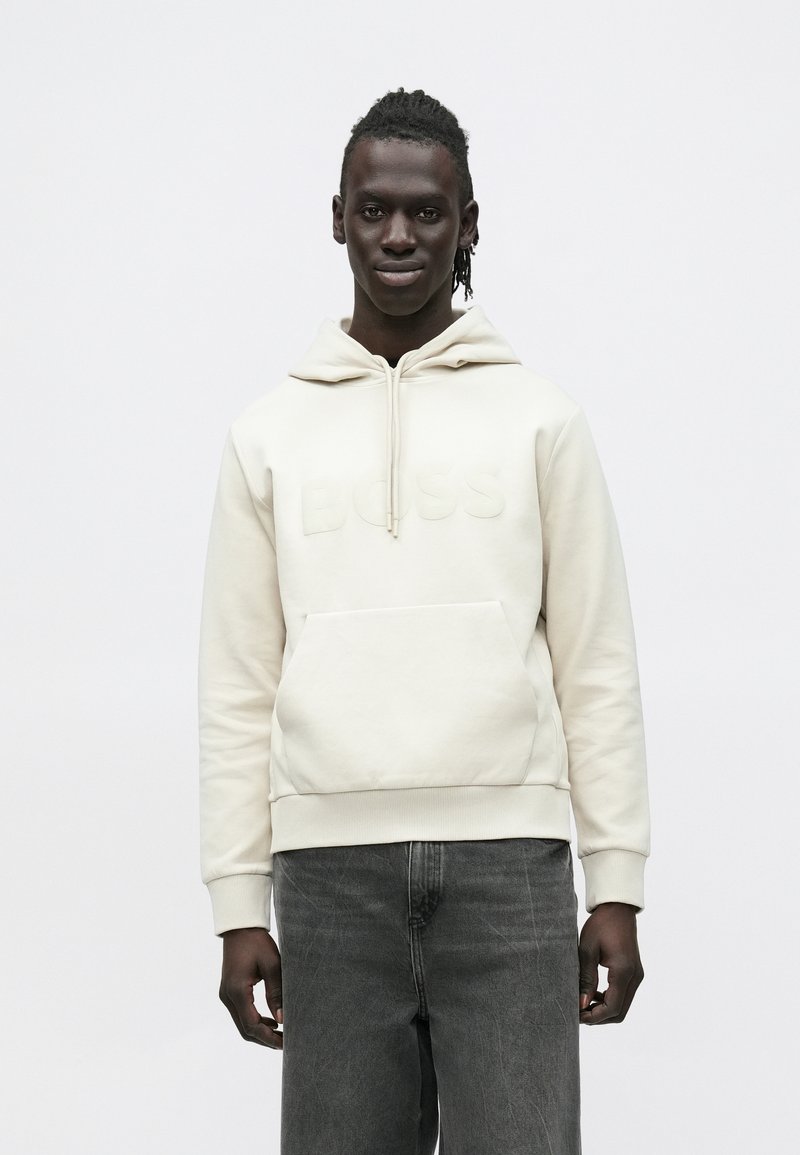 Νεαρός άνδρας με σκουρόχρωμο δέρμα και πλεγμένα μαλλιά, που φοράει ανοιχτό μπεζ φούτερ με κουκούλα με την επιγραφή "BOSS" και σκούρα γκρι τζιν, στέκεται μπροστά σε ένα απλό φόντο.