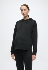 Czarna bluza z miękkiego materiału z kapturem na sznurki, luźnym krojem, długimi rękawami i bocznymi rozcięciami, zestawiona z dopasowanymi czarnymi spodniami.