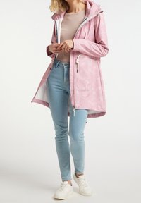 Veste imperméable rose pâle avec capuche, ornée de motifs floraux blancs, poches zippées et cordons ajustables. Associée à un jean bleu clair.