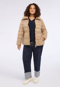 Beige pufferjacka med dragkedja, som har två framfickor och krage, i kombination med mörka jeans och ljusa sneakers.