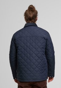 Marineblaue gesteppte Jacke mit Kragen, die ein Rautenmuster, sichtbare Nähte und Druckknöpfe an den Manschetten aufweist. Rückansicht dargestellt.