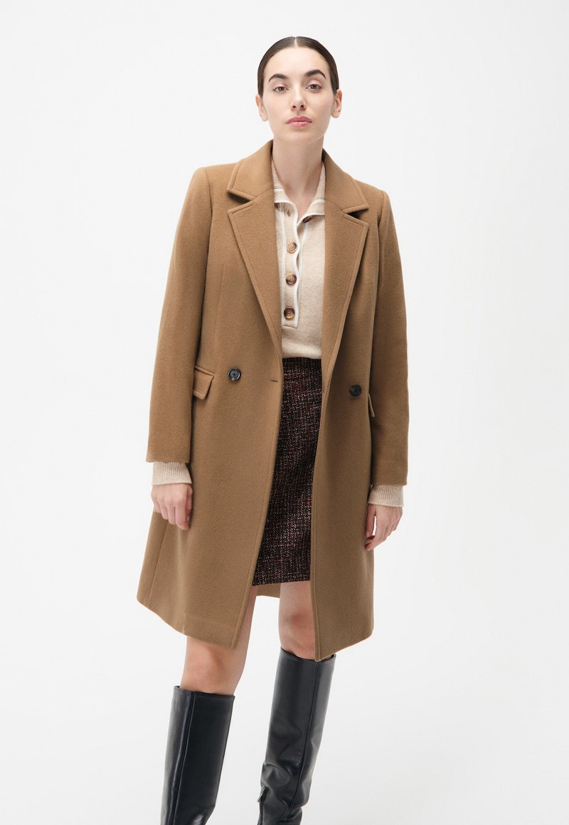 Bruine wollen jas van een wollen mix met ingesneden revers, dubbele knopen en zijzakken, gedragen over een beige blouse met knopen en een gestructureerde donkere rok.
