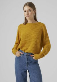 Gul stickad tröja med ett texturerat diamantmönster, bred halsringning och lösa ärmar, i kombination med högmidjade blå denimjeans.
