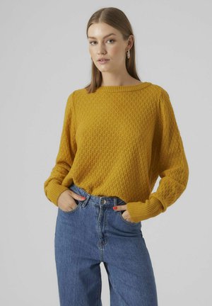 Gul strikket sweater med et struktureret diamantmønster, bred udskæring og løse ærmer, parret med højtaljede blå denimjeans.