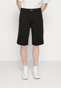 Muž v černých kraťasech do kolen, bílých ponožkách a bílých teniskách, stojící před jednoduchým bílým pozadím s rukama za zády.