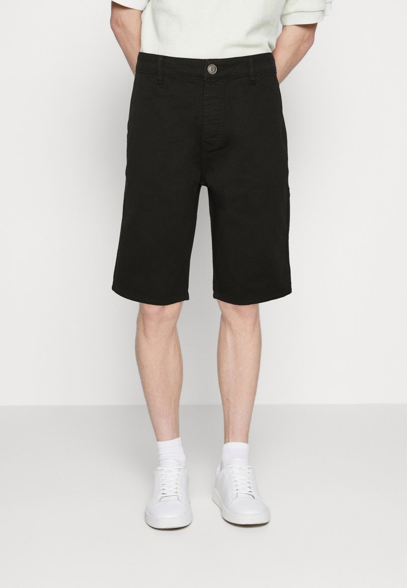 Muž v černých kraťasech do kolen, bílých ponožkách a bílých teniskách, stojící před jednoduchým bílým pozadím s rukama za zády.