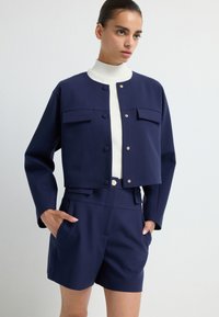 Granatowa krótka kurtka z dwoma kieszeniami na piersi, złotymi dodatkami i białym swetrem z wysokim kołnierzem, zestawiona z dopasowanymi granatowymi szortami. Gładka faktura.